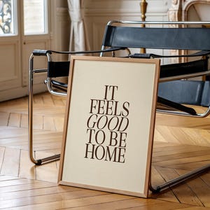 May include: A framed print with the text "IT FEELS GOOD TO BE HOME" in brown letters. The frame is light brown, and the background is a cream color. The print is leaning against a chair with a black seat and chrome frame.