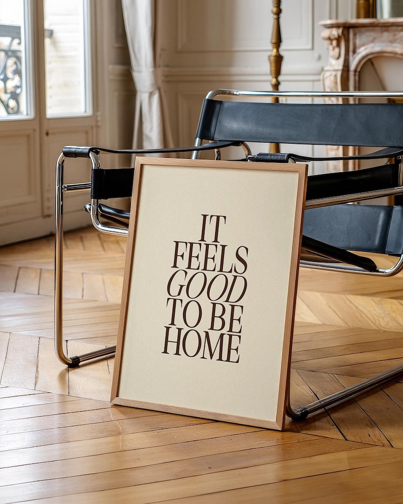 May include: A framed print with the text "IT FEELS GOOD TO BE HOME" in brown letters. The frame is light brown, and the background is a cream color. The print is leaning against a chair with a black seat and chrome frame.