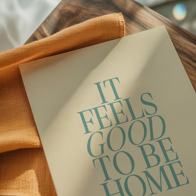 May include: A rectangular print with the text "IT FEELS GOOD TO BE HOME" in teal lettering. The print is on a cream-colored background, with a folded orange cloth and a wooden surface in the background.