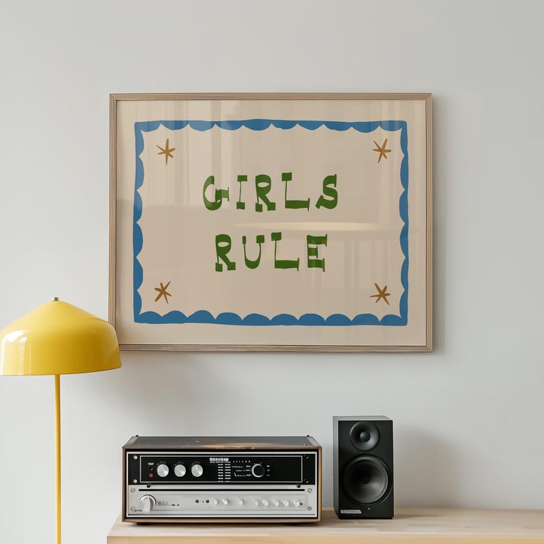 May include: A framed art print with the words "GIRLS RULE" in green lettering. The print has a blue scalloped border and star accents. A yellow lamp, stereo equipment, and a black speaker are below the artwork.