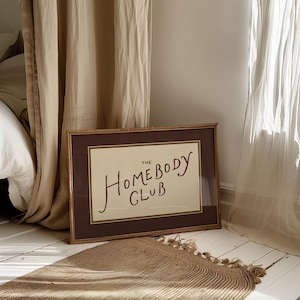 May include: A framed print with the text "The Homebody Club" in a handwritten style. The frame is brown with a woven texture, and the print has a cream-colored background. A woven rug and curtains are in the background.