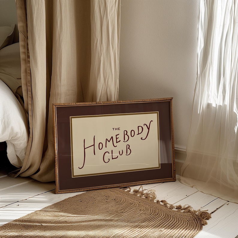 May include: A framed print with the text "The Homebody Club" in a handwritten style. The frame is brown with a woven texture, and the print has a cream-colored background. A woven rug and curtains are in the background.