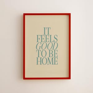 May include: A framed print with a red border. The print has a cream-colored background with the words "IT FEELS GOOD TO BE HOME" in teal-colored, stylized text. The frame is rectangular and the print is centered.