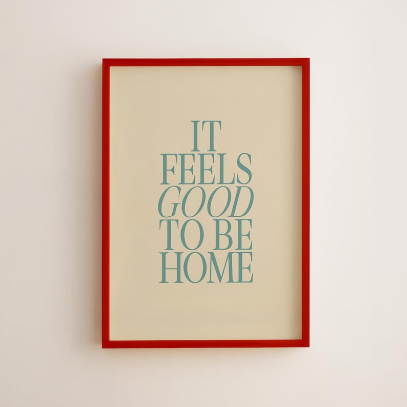 May include: A framed print with a red border. The print has a cream-colored background with the words "IT FEELS GOOD TO BE HOME" in teal-colored, stylized text. The frame is rectangular and the print is centered.