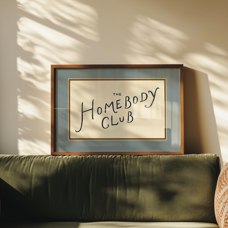 May include: A framed print with the text "THE HOMEBODY CLUB" in a handwritten style. The artwork is set against a light beige background, framed in a wooden frame with a blue border. The print rests on a green velvet sofa.