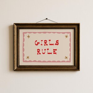 May include: A framed print with the words "GIRLS RULE" in red, centered on a cream background. The print is bordered by a pink scalloped edge and adorned with four gold star accents. The frame is a dark, ornate gold.