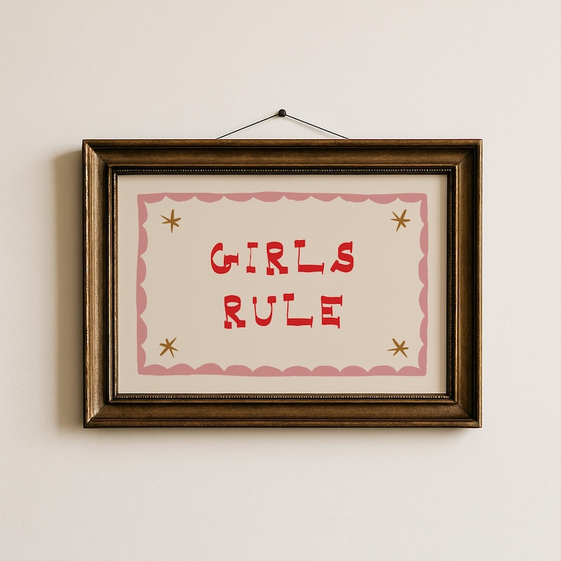 May include: A framed print with the words "GIRLS RULE" in red, centered on a cream background. The print is bordered by a pink scalloped edge and adorned with four gold star accents. The frame is a dark, ornate gold.
