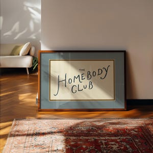 May include: A framed art print with the text "THE HOMEBODY CLUB" in a handwritten style. The artwork is set against a cream background, framed with a blue border and a brown wooden frame. The print is displayed on a wooden floor.