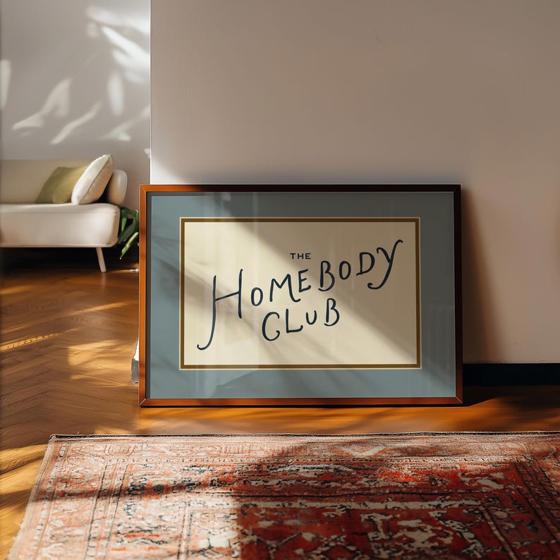 May include: A framed art print with the text "THE HOMEBODY CLUB" in a handwritten style. The artwork is set against a cream background, framed with a blue border and a brown wooden frame. The print is displayed on a wooden floor.