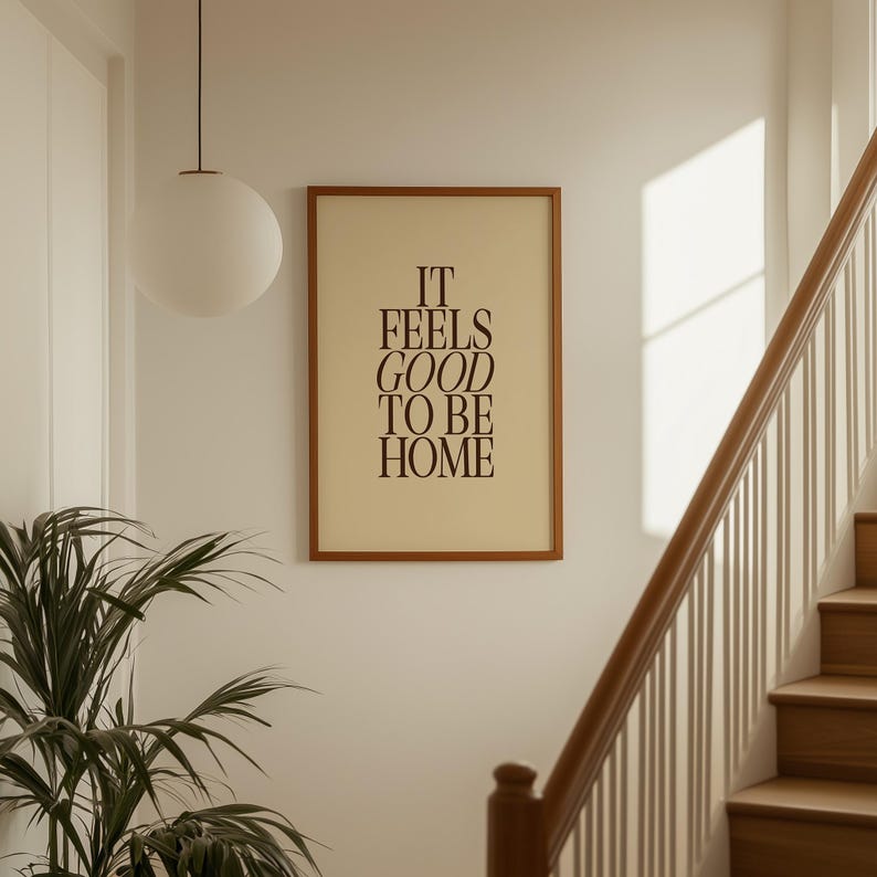 May include: A framed print with the text "IT FEELS GOOD TO BE HOME" in a brown serif font. The print is in a brown wooden frame and hangs on a white wall. A white globe light hangs from the ceiling, and a staircase is visible.