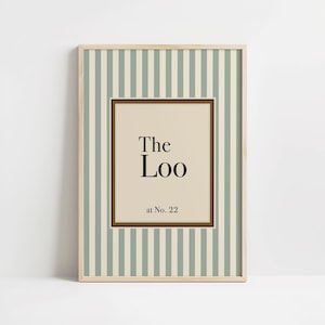 May include: Framed wall art with a light wood frame. The artwork features a striped background in white and pale green. The center has a beige rectangle with the words "The Loo" and "at No. 22" in black text.