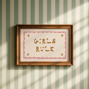 May include: A framed print with the words "GIRLS RULE" in brown letters on a cream background. The print has a pink scalloped border and star accents. The frame is a warm brown color, and the background is a striped pattern of white and light green.