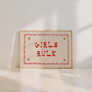 May include: A framed print with the words "GIRLS RULE" in red, stylized lettering. The text is centered on a cream-colored background, framed by a pink scalloped border and gold star accents. The frame is a light wood color.