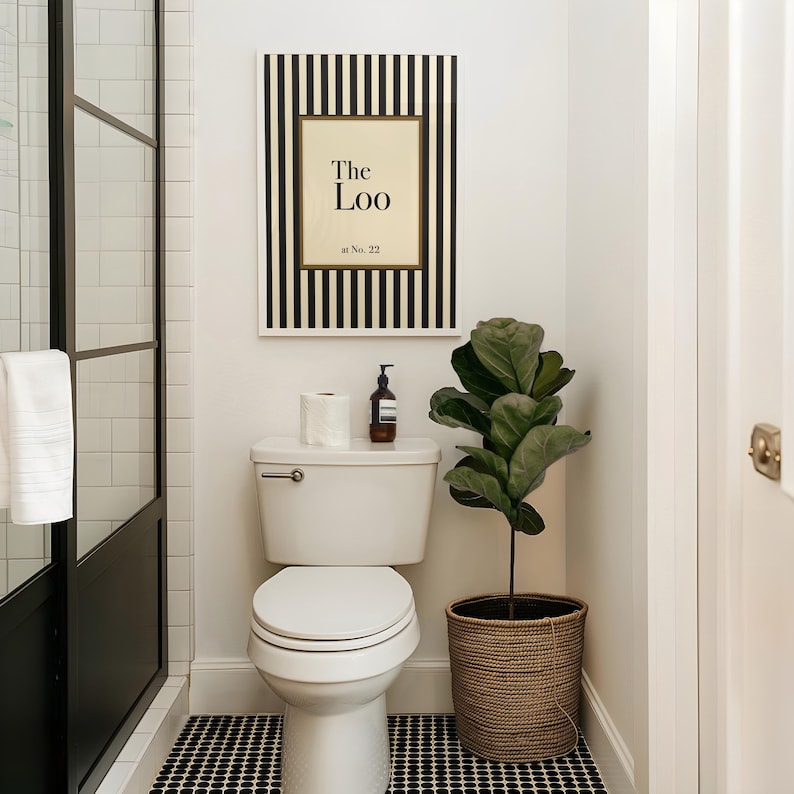May include: A modern bathroom scene featuring a white toilet, a framed sign that reads "The Loo", and a potted plant. The sign has a black and white striped border. The floor has a black and white tile pattern.