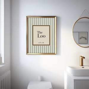 May include: A framed print with the text "The Loo" in a bathroom setting. The print has a gold frame and a striped background in light green and white. A round mirror and a white sink are also visible.