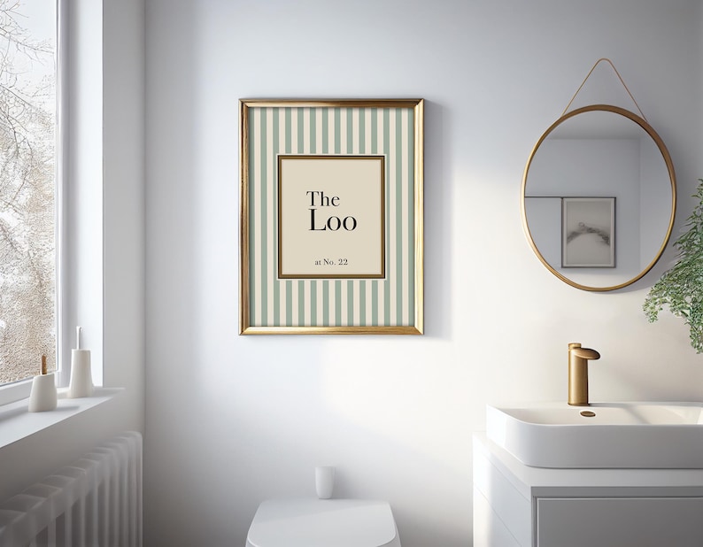 May include: A framed print with the text "The Loo" in a bathroom setting. The print has a gold frame and a striped background in light green and white. A round mirror and a white sink are also visible.