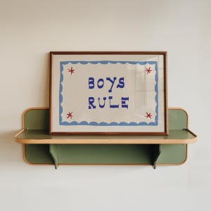 May include: A framed print with the words "BOYS RULE" in blue letters, surrounded by a blue scalloped border and red star accents. The print is displayed on a green and wood-toned shelf.