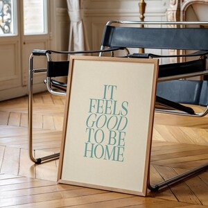 May include: A framed print with the text "IT FEELS GOOD TO BE HOME" in teal lettering. The print has a light beige background and a natural wood frame. The print is leaning against a black leather chair.