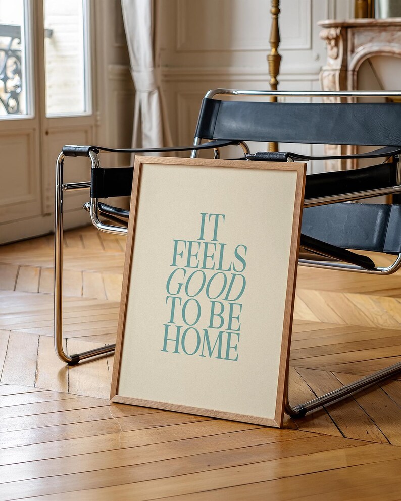 May include: A framed print with the text "IT FEELS GOOD TO BE HOME" in teal lettering. The print has a light beige background and a natural wood frame. The print is leaning against a black leather chair.