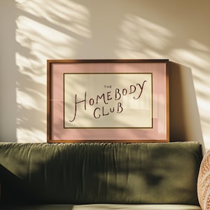 May include: A framed print with the words "THE HOMEBODY CLUB" in a handwritten style. The print has a pink border and a gold inner frame, set against a cream background. The frame is brown and the print is displayed above a green velvet sofa.