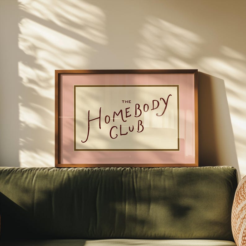 May include: A framed print with the words "THE HOMEBODY CLUB" in a handwritten style. The print has a pink border and a gold inner frame, set against a cream background. The frame is brown and the print is displayed above a green velvet sofa.
