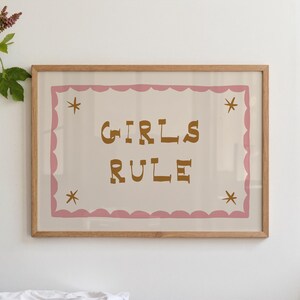 May include: A framed print with the words "GIRLS RULE" in brown letters. The print has a pink scalloped border and four star accents. The frame is made of light brown wood.
