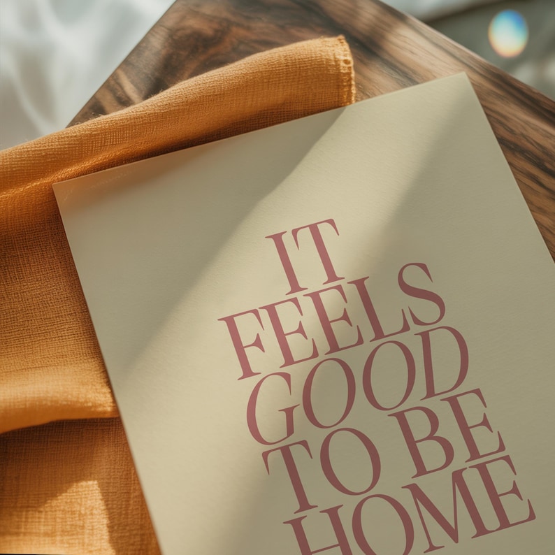 May include: A rectangular print with the text "IT FEELS GOOD TO BE HOME" in a burgundy color. The print is on a cream-colored background, with a folded orange fabric and a wooden surface in the background.