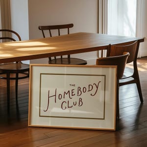 May include: A framed print with the text "THE HOMEBODY CLUB" in a handwritten style. The print is in a light wood frame and is leaning against a wooden floor. A wooden table and chairs are in the background.