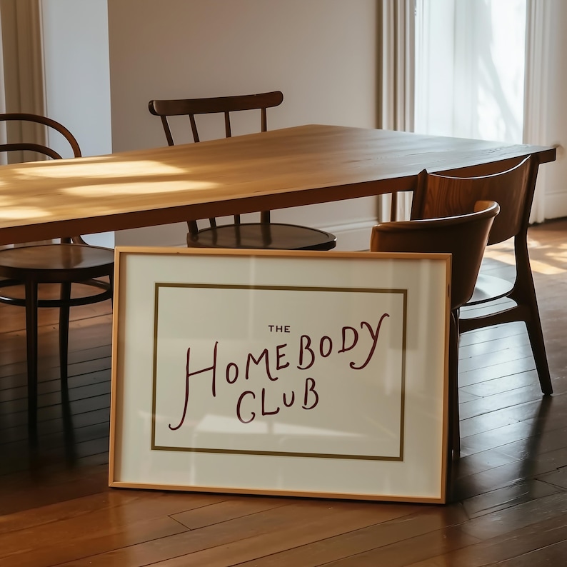 May include: A framed print with the text "THE HOMEBODY CLUB" in a handwritten style. The print is in a light wood frame and is leaning against a wooden floor. A wooden table and chairs are in the background.