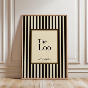 May include: Framed wall art with a cream-colored background and the text "The Loo at The Gables." The artwork is framed in a wooden frame and features a black and cream striped border.