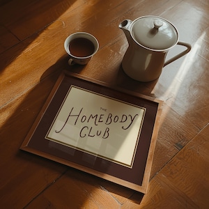 May include: A framed print with the text "The Homebody Club" sits on a wooden floor, next to a cup of tea and a beige teapot. The print has a brown frame and a cream-colored background. The teapot has a lid and a handle.