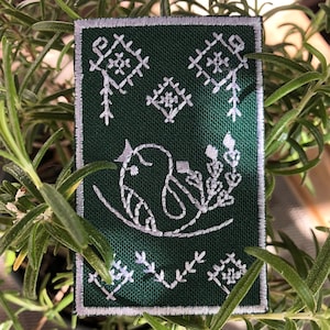 Puede incluir: Un parche rectangular bordado en verde oscuro con un borde blanco. El parche presenta un diseño de pájaro blanco posado en una rama, rodeado de patrones florales y geométricos. El parche está rodeado de hojas verdes.