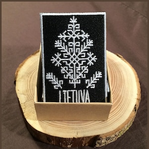 Pode incluir: Um remendo bordado preto com um design de árvore estilizado branco e a palavra "LIETUVA" abaixo. O remendo é exibido em uma superfície de madeira.