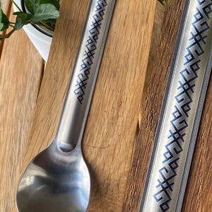 Op de afbeelding: Twee zilveren lepels met een blauw en wit geometrisch patroon op de steel. De lepels liggen op een houten oppervlak.