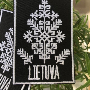 Può includere: Una toppa ricamata nera con un designo stilizzato di albero bianco e la parola "LIETUVA" sotto.