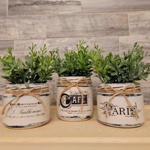 May include: Three decorative white jars with green plants, each featuring a different French-inspired design and text. The jars are tied with twine and sit on a wooden surface. The background is a gray brick wall.