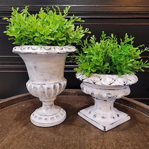May include: Two white urn-shaped planters with a distressed finish and dark grey accents, each containing a vibrant green artificial plant. The taller planter features a fluted base and bowl, while the shorter one has a square base.