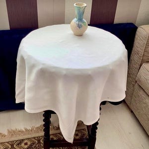May include: A small, round, dark wood table with a white tablecloth. A small, decorative vase with a light blue and beige floral design sits on top. The table is approximately 61 cm in diameter.