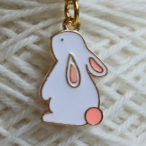 May include: A gold-colored metal stitch marker with a white bunny charm. The bunny has pink accents on its ears and tail. The stitch marker is attached to a gold-colored clasp.