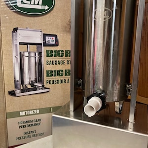 Puede incluir: Un embutidor de salchichas motorizado plateado con una boquilla de plástico blanco. El embutidor está etiquetado con "BIG BOY SAUSAGE STUFFERS" y "BIG BOY POUSSOIR A SAUCISSE". El embutidor está sobre una superficie blanca con una caja detrás.