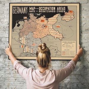Puede incluir: Un mapa vintage enmarcado de Alemania, que representa las zonas de ocupación. El mapa presenta texto en inglés y alemán, con varias banderas que marcan diferentes regiones. El mapa es sostenido por una persona contra una pared de ladrillos texturizada.
