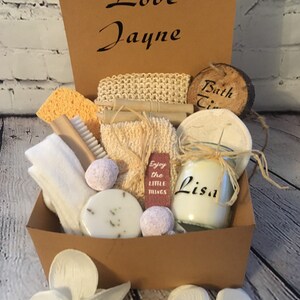 May include: A brown gift box containing bath and spa items. It features a candle, bath bombs, soap, a loofah, a brush, and a sponge. The box is adorned with the words "Love Jayne" and "Lisa". Two white orchids are in the foreground.