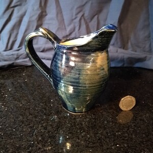 May include: A blue and green ceramic pitcher with a wide mouth and a handle. The pitcher has a glossy finish and is decorated with streaks of blue and green glaze.