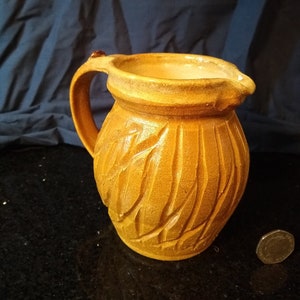 May include: A small, yellow ceramic jug with a textured, leaf-like design. The jug has a handle and a wide mouth.