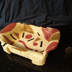 May include: A rectangular ceramic dish with a white base and a colorful geometric design. The design features red, brown, and black shapes on a white background.