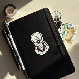 May include: A black journal with a black elastic band, featuring insect-themed stickers and black pens. A roll of gray washi tape sits at the top left. The stickers include a beetle and two frogs. The journal is closed.