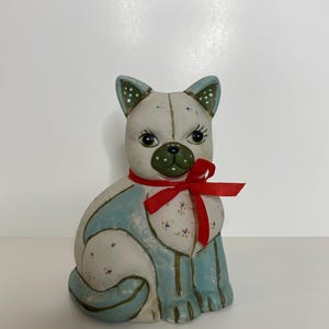 May include: A ceramic cat figurine with a white body, blue and green painted details, and a red bow around its neck.