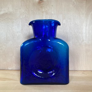 May include: A cobalt blue glass pitcher with a textured surface. The pitcher has a wide mouth and a square body.