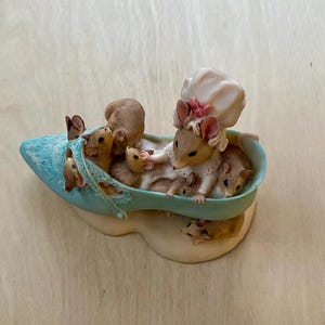 May include: A blue shoe figurine with a mother mouse and her baby mice inside. The mother mouse is wearing a white bonnet and a pink bow. The baby mice are all brown and white.