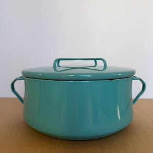 May include: A teal enamel pot with a matching lid. The pot has two side handles and a single handle on the lid. The pot is sitting on a wooden surface. Kitchenware.